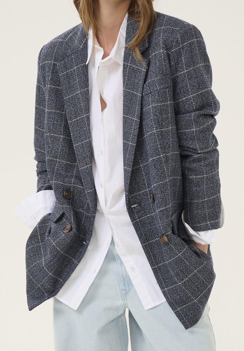 Doppelreihiger Blazer in Marineblau und Weiß mit Karomuster, strukturierter Stoff, mit großen Knöpfen, kombiniert mit einem weißen Hemd und hellblauen Jeans.