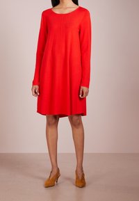 Abito rosso a maniche lunghe con scollatura rotonda, tessuto leggero, forma ad A, dettaglio della cucitura lungo la parte frontale, abbinato a scarpe con tacco color beige.