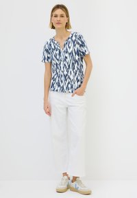 Top a maniche corte in blu e bianco con motivo astratto, caratterizzato da scollatura a V. Abbinato a pantaloni bianchi a gamba larga e sneakers beige.