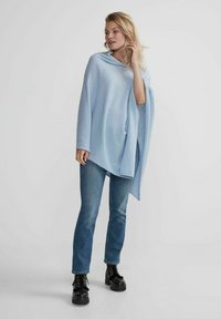 Ljusblå poncho med draperad design, med vid halsringning och sidoflikar, parat med blå jeans och svarta klumpiga stövlar.