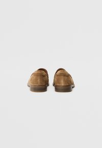 Paire de mocassins en daim marron avec petits talons en bois, vus de dos sur un fond blanc uni.