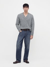 Homme portant un pull gris à col en V, un jean bleu ample avec une ceinture marron et des chaussures en cuir marron foncé, debout devant un fond blanc uni.