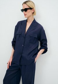 Blusa azul navy, con texture, colletto, due tasche sul petto e maniche arrotolate. Abbinata a pantaloni larghi coordinati.