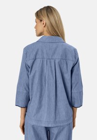 Camicia di denim azzurro chiaro con maniche a tre quarti, colletto classico e orlo con dettagli a scallop. Caratterizzata da una vestibilità rilassata e pieghe sul retro.