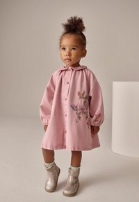 Abito di cotone rosa con colletto volant, bottoni frontali e ricamo floreale raffigurante una farfalla. Il bambino indossa stivali metallici argento.