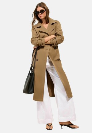 Žena v béžovom trenčkotku, biele široké nohavice, čierne sandále na podpätku, čierna kabelka a tmavé slnečné okuliare so skríženými rukami.