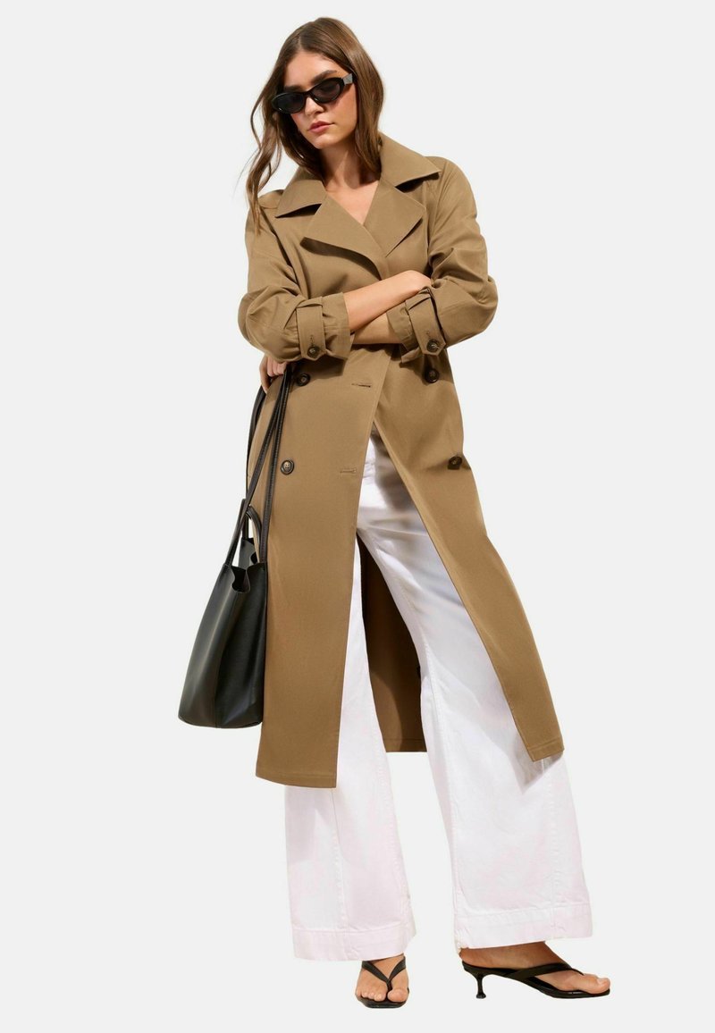 Kvinde iført beige trenchcoat, hvide bukser med vide ben, sorte sandaler med hæl, sort håndtaske og mørke solbriller med armene krydset.