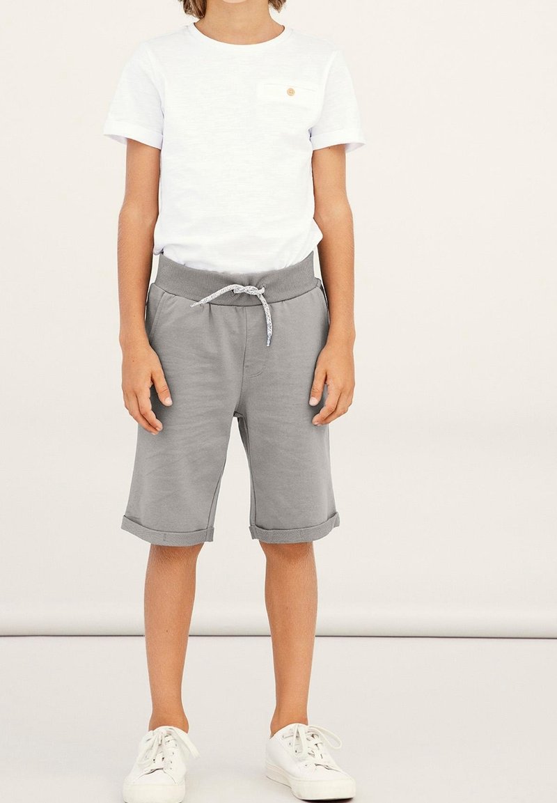 Enfant portant une chemise blanche à manches courtes avec poche et un short gris jusqu'aux genoux avec cordon, debout, les bras détendus.