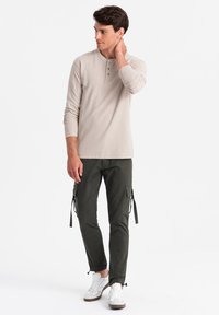 Lichtbeige langemouwen henley shirt met drie knopen; gecombineerd met olijfgroen cargobroek met zakken en verstelbare koordjes. Witte sneakers.