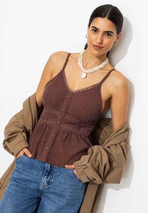 Femme aux cheveux foncés vêtue d'un haut marron texturé sans manches, d'une veste beige posée sur les épaules, d'un jean bleu, et d'un collier blanc à perles avec un pendentif rond.