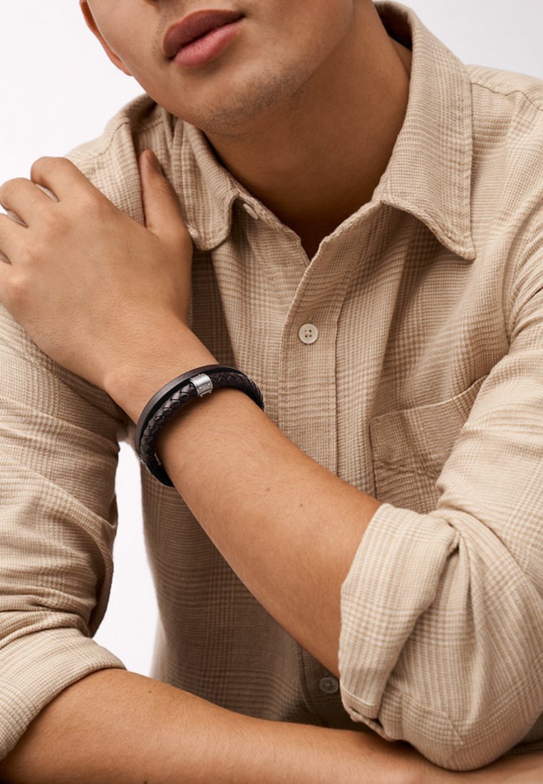 Schwarzes geflochtenes Lederarmband mit einem silberfarbenen Verschluss, getragen am Handgelenk, kontrastiert mit einem hellen Karoshirt.