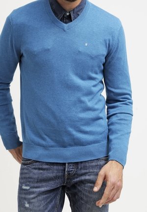 Homme portant un pull bleu clair à col en V par-dessus une chemise sombre boutonnée et un jean bleu déchiré, sur un fond blanc uni.