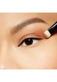 Close-up van een oog, met een gladde bruine ooglid en gedefinieerde zwarte eyeliner, terwijl een make-upkwast product aanbrengt bij de buitenste hoek.