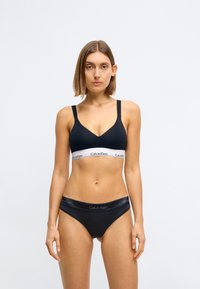 Schwarzer Crop-BH mit breiten Trägern und einem weißen elastischen Bund, der das Calvin Klein-Logo trägt. Passende schwarze Bikinihöschen mit Logo-Bund.