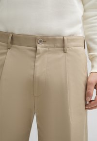 Pantaloni beige con una texture liscia, dotati di chiusura con bottone, passanti per cintura e una tasca frontale singola. Tessuto resistente alle pieghe.