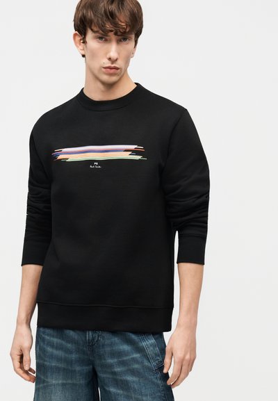 Νέος άνδρας με μαύρο φούτερ που έχει σχεδιασμό με πολύχρωμες οριζόντιες βουρτσιές και μπλε τζιν, στέκεται μπροστά σε απλό φόντο.