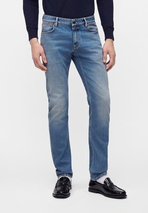 Personne portant un jean slim bleu clair, des mocassins en cuir noir, des chaussettes blanches et un haut à manches longues foncé, debout devant un fond uni.