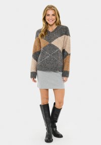 V-Ausschnitt, Übergrößenpullover mit einem Strickmuster in grau, braun und cremefarbenen Rauten. Kombiniert mit einem grauen Rock und schwarzen Stiefeln bis zum Knie.
