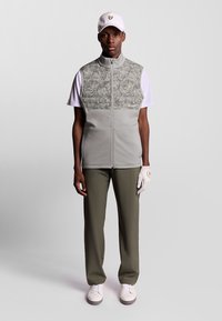 Grå vest med tekstureret grøn camouflagepanel, hvid T-shirt, olivengrønne bukser, golfhandske og hvide sko; vesten har et logo.