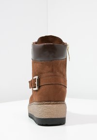 Brązowy zamszowy botek za kostkę z brązowym skórzanym mankietem, bocznym zamkiem błyskawicznym i ozdobną metalową klamrą. Gruba, teksturowana podeszwa zapewnia dodatkową przyczepność.