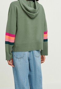 Felpa con cappuccio verde oliva con orlo a coste. Una manica presenta audaci strisce orizzontali in rosa, arancione e verde. Indossata con jeans azzurri.