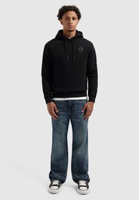 Jonge man die staat, gekleed in een zwarte hoodie met klein borstlogo, vervaagde spijkerbroeken met brede pijpen in het blauw, en zwart-witte sneakers, op een witte achtergrond.