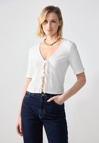 Crop top blanc texturé avec manches courtes, décollet�é en V profond et détail lacé à l'avant. Porté avec un jean bleu foncé taille haute. Collier épais en or.