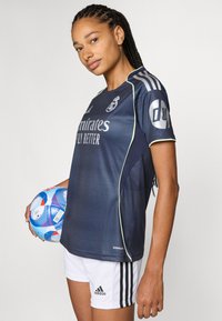 Dark blue soccer jersey with light green accents, featuring the Real Madrid logo and "Emirates Fly Better" text. Paired with white shorts.