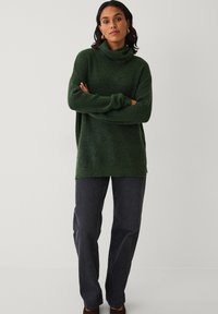 Grøn strikket rullekravesweater med løs pasform, lange ærmer og ribbet struktur, parret med mørke jeans og brune sko.