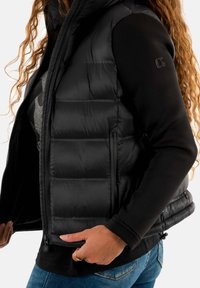 Gilet matelassé noir avec col montant, fermeture éclair sur le devant et poches latérales. Porté par-dessus un haut noir, avec un tissu texturé et une coupe ajustée.