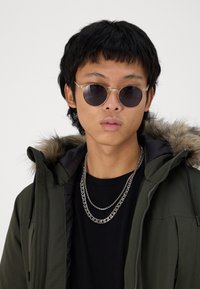 Green hooded jacket with fur trim, worn over a black shirt. Silver chains visible. Clear, round sunglasses with dark lenses. Short, textured hair.