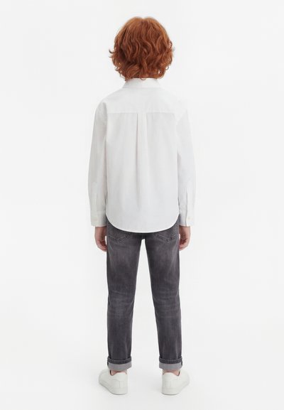 Enfant aux cheveux roux bouclés, de dos, portant une chemise blanche à manches longues, un jean foncé retroussé et des baskets blanches sur un fond blanc.