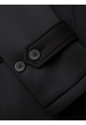 Primo piano della manica di un cappotto nero che mostra due bottoni neri e una cucitura dettagliata sul polsino in tessuto spesso.