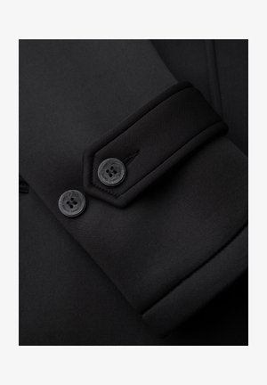 Primo piano della manica di un cappotto nero che mostra due bottoni neri e una cucitura dettagliata sul polsino in tessuto spesso.