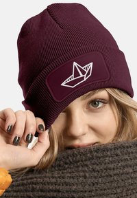 Burgundy knit beanie with a textured surface and a contrast patch featuring a white origami boat design. Soft material, snug fit.