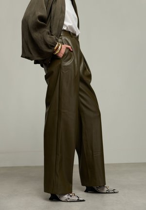 Femme portant un pantalon ample en cuir olive, une veste assortie à manches bouffantes, un haut blanc, des bracelets dorés et des escarpins pointus en peau de serpent.