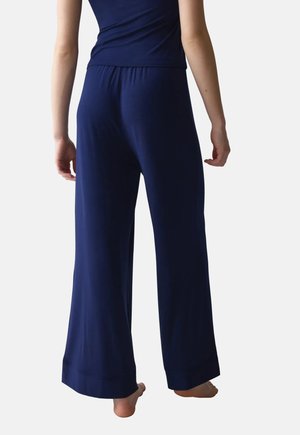 Personne portant un pantalon bleu marine à jambes larges et un haut assorti, debout pieds nus devant un fond clair uni.