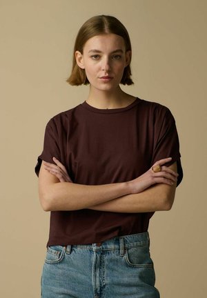 Jonge vrouw met kort bruin haar staat met gekruiste armen, draagt een donkerbruine T-shirt en blauwe spijkerbroek, tegen een beige achtergrond.