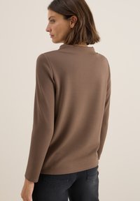 Bruine lange mouwen top gemaakt van zachte stof. Heeft een subtiele kraag, rechte snit en een soepele textuur, gecombineerd met donkere broek.