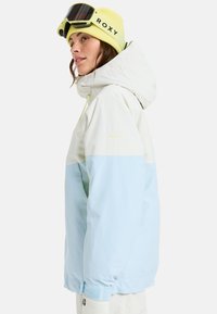 Color-blocked snow jacket in white and light blue, featuring a hood, adjustable drawstring, and Roxy branding. Complements with a yellow beanie.