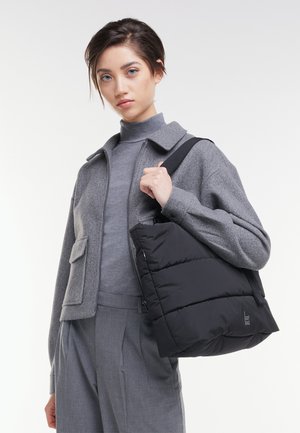 Jeune personne portant une tenue grise tient un grand sac à bandoulière noir matelassé avec une étiquette "GOT BAG" sur fond blanc.