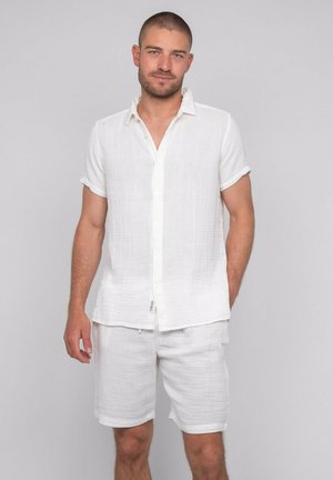 Jeune homme aux cheveux courts portant une chemise blanche à manches courtes boutonnée et un short blanc assorti sur un fond uni.