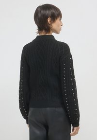 Frau mit kurzen Haaren, die einen schwarzen Strickpullover mit strukturierten Mustern und kleinen silbernen Nieten an den Ärmeln trägt, kombiniert mit schwarzen Lederhosen.
