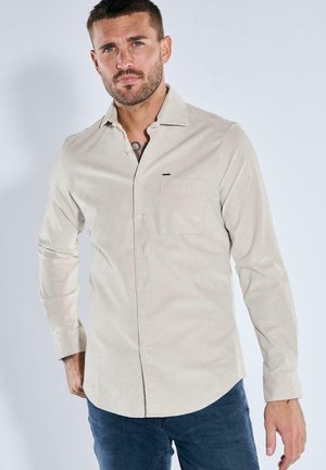 Mann mit kurzen Haaren und Bart, der ein beigefarbenes Hemd mit Knöpfen und blaue Jeans trägt, vor einem schlichten hellen Hintergrund stehend.