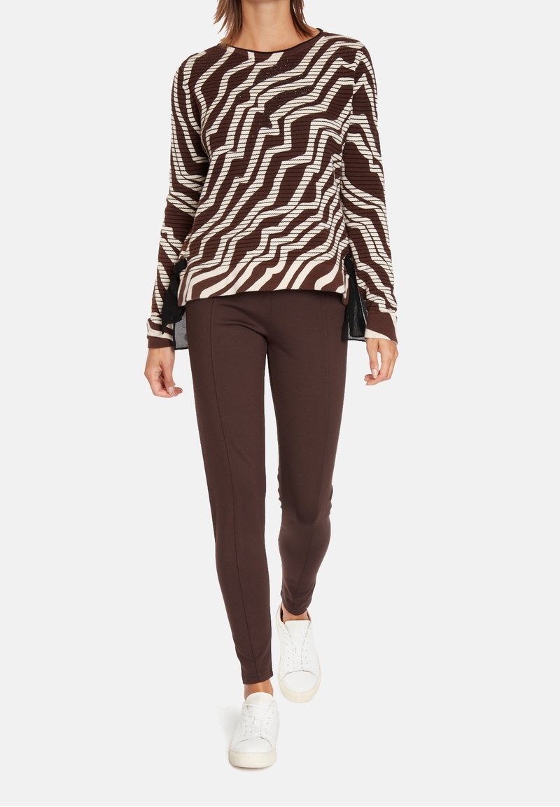 Brun och krämfärgad zigzag-mönstrad stickad tröja i kombination med bruna tajta byxor och vita sneakers. Tröjan har sidoflikar för en extra design.