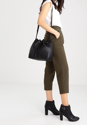 Kvinde iført olivengrønne korte bukser, hvid ærmeløs top, sorte ankelstøvler med hæl, bærende en sort læder bøtte taske på skulderen.