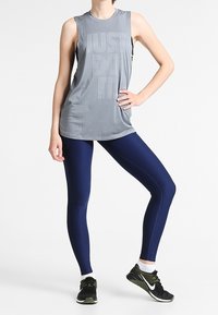 Grijze mouwloze sporttop met ribbelstructuur en "JUST DO IT" bedrukking; gecombineerd met marineblauwe leggings en zwarte sportschoenen.