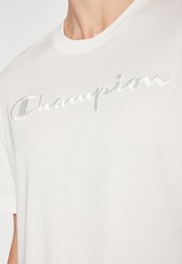 Weißes Baumwoll-T-Shirt mit einem silbernen, gestickten "Champion"-Logo auf der Brust. Kurze Ärmel und ein runder Ausschnitt.