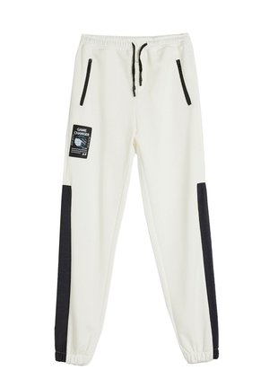 Pantalones de chándal blancos con paneles laterales negros, cinturilla elástica, cordón y dos bolsillos con cremallera. Presenta una etiqueta "GAME CHANGER" en la parte frontal.