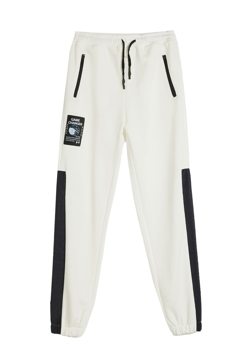 Pantalones de chándal blancos con paneles laterales negros, cinturilla elástica, cordón y dos bolsillos con cremallera. Presenta una etiqueta "GAME CHANGER" en la parte frontal.